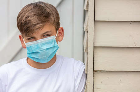 Boy teenager teen male child wearing a face mask outside leaning against the side of a house during the Coronavirus COVID-19 virus  pandemicの写真素材