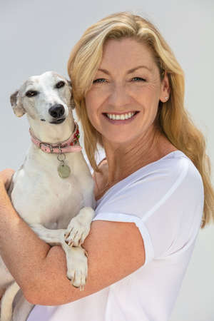 Portrait of an attractive middle aged blonde woman smiling with perfect teeth holding her pet whippet dogの写真素材