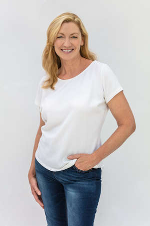 Studio portrait of an attractive middle aged blonde woman smiling on a white background wearing a white t-shirt and blue jeansの写真素材