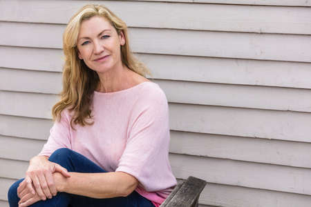 Outdoor portrait of an attractive middle aged blonde woman smiling sitting on a wooden garden chairの写真素材
