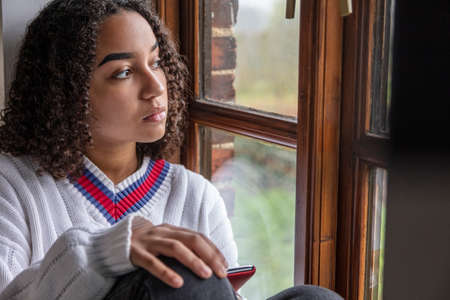 Beautiful mixed race African American girl teenager female young woman sad depressed or thoughtful looking out of a window using mobile cell phoneの写真素材