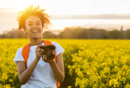 Beautiful mixed race African American girl teenager female young woman taking photographs with a camera at sunset or sunrise in a field of yellow flowersの写真素材