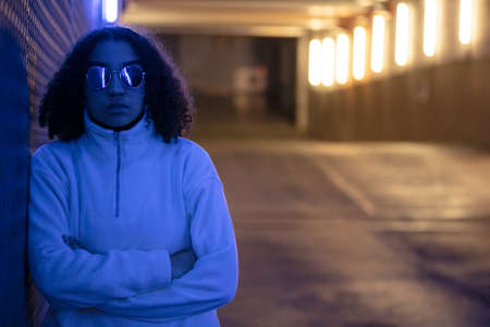 Beautiful mixed race African American girl teenager female young woman wearing sunglasses in a modern blue underground urban city environmentの写真素材