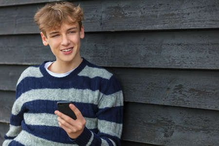 Smiling Happy male boy teenager outside leaning wall using his mobile cell phone for social media or video callの写真素材