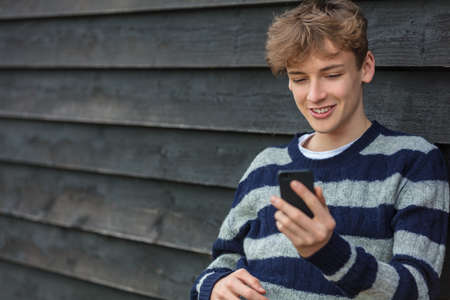 Smiling Happy male boy teenager outside leaning wall using his mobile cell phone for social media or video callの写真素材