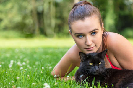 Beautiful female teenager girl young woman with blue eyes, laying down outside on grass with a black catの写真素材