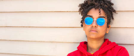 Panorama girl teenager cool teen mixed race biracial African American female young woman wearing blue sunglasses and a red hoodie on vacation in summer sunshine panoramic web bannerの写真素材
