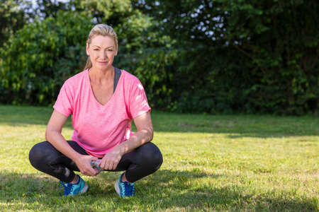Outdoor portrait of an attractive middle aged blonde woman smiling drinking water relaxing after exercising, running or fitness lifestyleの写真素材