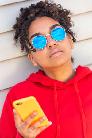 Girl teenager cool teen mixed race biracial African American female young woman wearing blue sunglasses and a red hoodie using cell phone on vacation in summer sunshineの写真素材