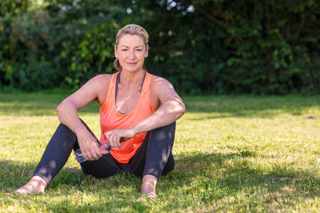 Outdoor portrait of an attractive middle aged blonde woman smiling drinking water relaxing after yoga, exercising, running or fitness lifestyleの写真素材