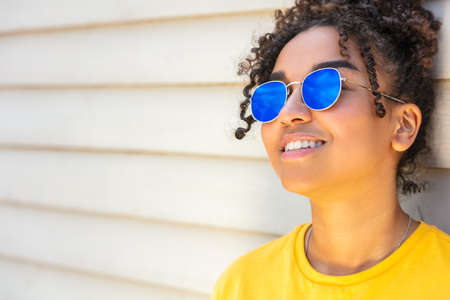 Girl teenager cool teen mixed race biracial African American female young woman wearing blue sunglasses and a yellow t-shirt on vacation smiling in summer sunshineの写真素材