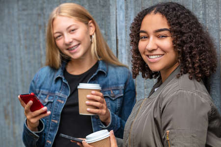 Interracial friends pretty blonde teenager and mixed race African American girl female young woman using their smart phones for social media and drinking takeout coffee outsideの写真素材
