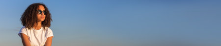 Panoramic web banner beautiful and thoughtful mixed race African American female girl child teenager young woman in sunshine wearing sunglasses with blue sky panoramaの写真素材