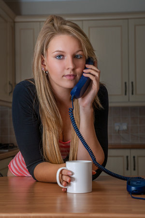 Young woman or teenager talking on a landline telephone at home in her kitchen drinking mug of coffee or teaの写真素材