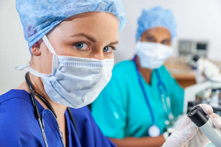Female medical or woman scientist weaing face mask using a microscope in a research lab or laboratory with her colleagueの写真素材