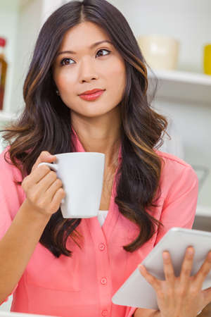 Beautiful Asian Chinese Woman using tablet computer for social media and drinking coffee or tea in her kitchen at homeの写真素材
