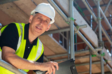 Male builder foreman, construction worker or site manager holding a clipboard, wearing a white hard hat and hi vis vestの写真素材