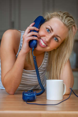 Happy smiling young woman or teenager talking on a landline telephone at home in her kitchen drinking mug of coffee or teaの写真素材