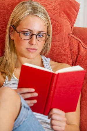Beautiful young woman teenager girl with blonde hair and blue eyes wearing glasses relaxing at home reading a red bookの写真素材