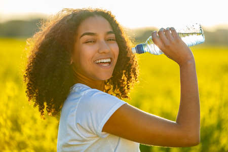 Outdoor portrait of beautiful happy mixed race African American girl teenager female young woman drinking water from a bottle in a field of yellow flowers at sunset in golden evening sunshineの写真素材