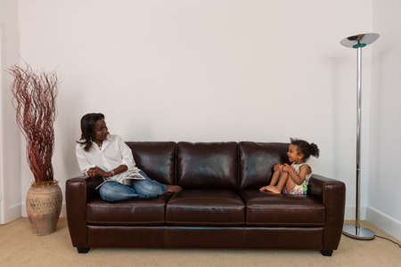 Happy African American black mother and her mixed race daughter, woman and girl child, laughing together at home on a leather sofa or setteeの写真素材