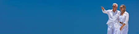 Panoramic happy senior man and woman couple walking together in bright clear blue sky, the man is pointing panorama web banner headerの写真素材
