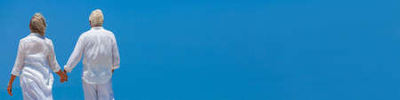 Panorama rear view of happy successful retired senior man and woman couple walking and holding hands with bright clear blue sky panoramic web bannerの写真素材