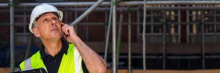 Panorama thoughtful male builder foreman, construction worker, contractor or architect on building site holding black clipboard and talking on his cell phone panoramic web banner headerの写真素材
