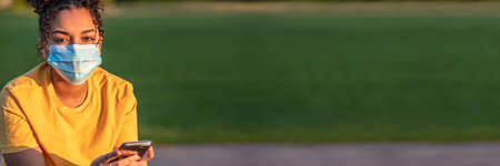 Panoramic web banner mixed race biracial African American teenager teen girl young woman wearing a face mask, using mobile cell phone during the pandemic, panorama headerの写真素材