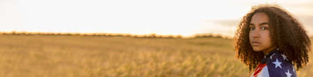 Panorama sad depressed mixed race African American girl teenager female young woman in a field of wheat or barley crops wrapped in USA stars and stripes flag in golden sunset evening sunshine panoramic web bannerの写真素材
