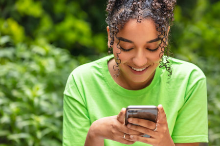 Outdoor portrait of beautiful happy mixed race African American girl teenager female young woman smiling using mobile cell phone for text messaging or social mediaの写真素材