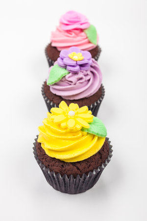 Cupcakes or cup cakes with icing or frosting, pink, yellow and purple flowers with green leaves photographed on a white backgroundの写真素材