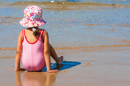 Rear view of cute little girl female child sitting playing on a beach on pink swimming costume and hatの写真素材
