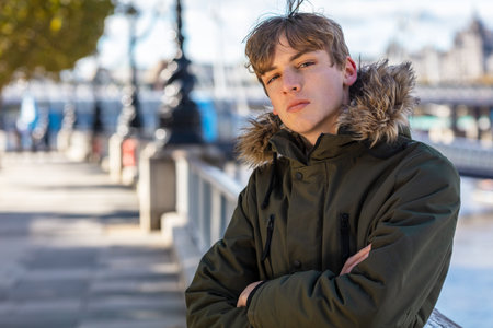 Young adult male boy teenager outside in sunshine wearing a hooded parka coat in an urban cityの写真素材