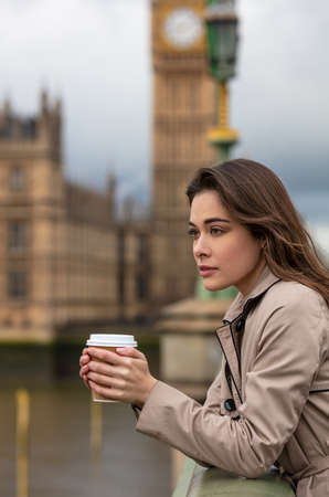 Beautiful sad, depressed or thoughtful young woman in London on Westminster Bridge over the River Thames drinking take out coffee by Big Benの写真素材
