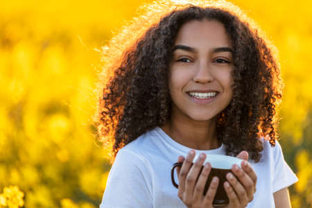 Beautiful happy mixed race African American girl teenager female young woman smiling drinking coffee or tea outdoors in a field of yellow flowersの写真素材