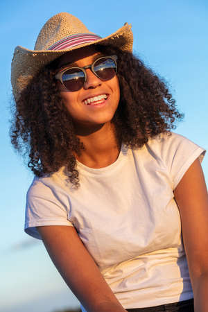 Beautiful and happy mixed race African American female girl teenager young woman with perfect teeth wearing straw cowboy hat sunglasses and white t-shirt in late evening sunshineの写真素材