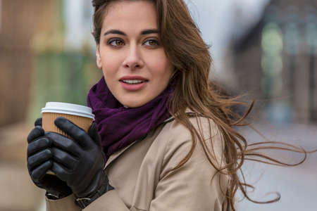 Girl or young woman drinking coffee or tea in a disposable cup outside wearing gloves, a scarf and raincoat in cold wet weatherの写真素材