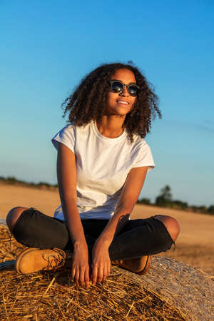 Beautiful mixed race African American female teenager young woman sitting on a hay bale in a field wearing white t-shirt and sunglasses at sunsetの写真素材