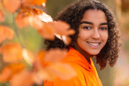 Outdoor portrait of beautiful happy mixed race biracial African American girl teenager female young woman smiling with perfect teeth wearing an orange hoodie surrounded by Fall or Autumn golden leavesの写真素材