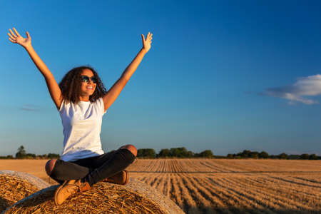 Beautiful, happy mixed race African American female girl child teenager young woman celebrating arms raised in sunshine wearing sunglasses and smiling with perfect teethの写真素材