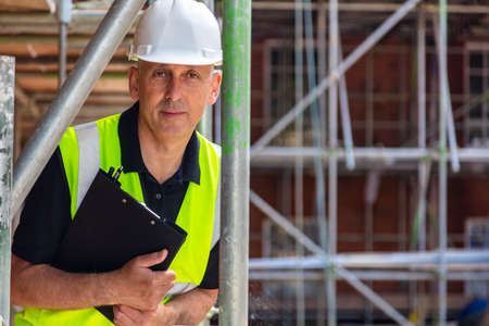 Serious looking male builder foreman, worker, contractor or architect on construction or building site holding black clipboard wearing white hard hatの写真素材