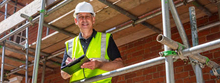 Panoramic web banner header male builder foreman, construction worker, contractor or architect on building site writing on black clipboard laughing, smiling and happy panoramaの写真素材