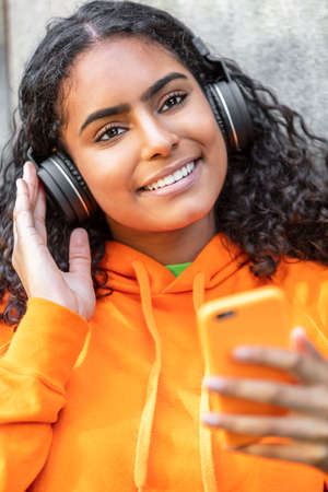 Outdoor portrait of beautiful happy mixed race African American girl teenager female young woman wearing an orange hoodie listening to music on wireless headphones and her mobile cell phoneの写真素材