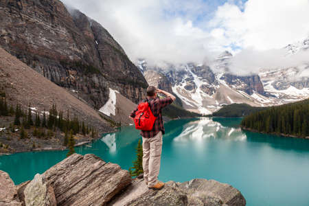 Hiking man with red rucksack backpack standing on a rock overlooking Moraine Lake looking at snow covered Rocky Mountain peaks, Banff National Park, Alberta Canadaの写真素材