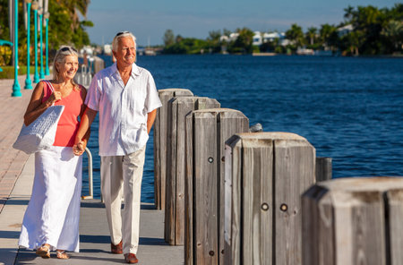 Happy senior man and woman romantic retired couple together holding hands looking out to tropical sea or river with bright clear blue skyの写真素材