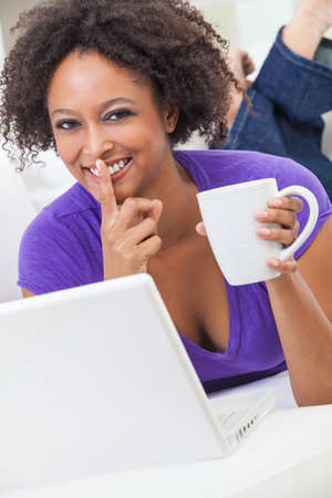 Beautiful happy mixed race African American girl or young woman laying down on sofa using a laptop computer for on line shopping or social media, finger to lips with secret and drinking tea or coffeeの写真素材