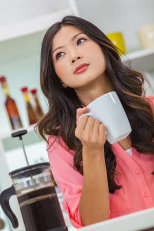 Beautiful young Asian Chinese woman or girl at home in her kitchen looking sad or thoughtful and drinking a cup or mug of french press coffeeの写真素材