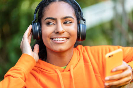 Outdoor portrait of beautiful happy mixed race African American girl teenager female young woman wearing an orange hoodie listening to music on wireless headphones and her mobile cell phoneの写真素材