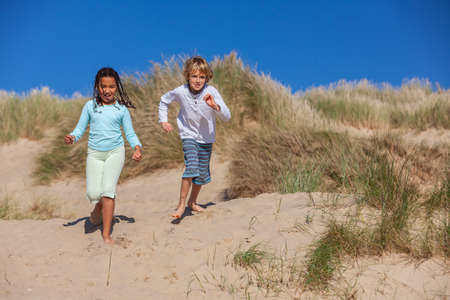 Two children, a blonde boy and a mixed race African American girl, having fun running racing in the dunes of a sandy beachの写真素材